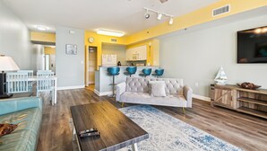 A modern living room with a sofa, coffee table, and a TV. There is a kitchen area with a bar and blue chairs.