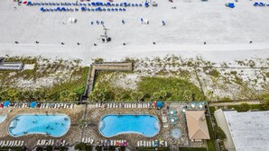 A beach with rows of blue chairs and umbrellas, a swimming pool area with multiple pools, and a building with a rooftop deck.
