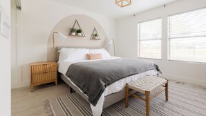 A bedroom with a bed, bedside tables, a bench, a hanging light fixture, and a window with blinds.