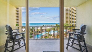 A balcony with two chairs, a pool, and a view of the beach and hotel buildings.
