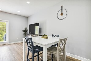 Un coin repas avec une table et des chaises blanches, un téléviseur accroché au mur et une plante posée sur la table.