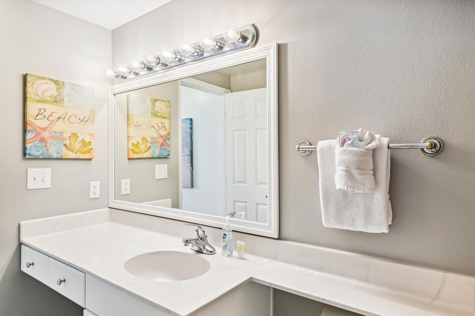 A bathroom with a white sink, a large mirror with lights, and a towel rack with a white towel.
