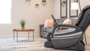 A modern living room with a massage chair, a grey sofa, a small side table with a plant, and a large mirror on the wall. - Kordeluxe Row #3 Studio Townhouse (Cedar City)