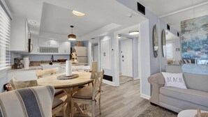 Una cocina moderna con armarios blancos, una isla central e iluminación colgante. Se aprecia un comedor con una mesa y sillas de madera. Se ve un salón con un sofá beige y un cuadro en la pared.