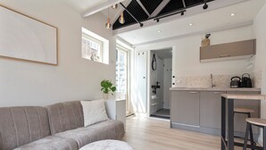 A modern kitchen with a grey countertop, a sink, and a bar stool. There is a grey sofa and a round ottoman in the living area. - The Roost - Cottage (Zandvoort)