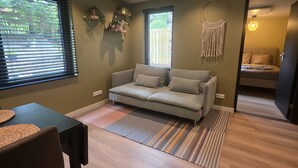 A modern living room with a grey sofa, a dining table with chairs, a macrame wall hanging, and a view of the outdoors through the windows. - Blissea - Comfortable stay, top location (Zandvoort)