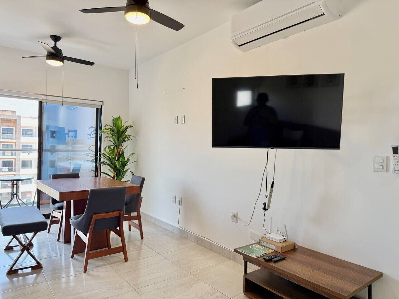A modern living room with a flat-screen TV mounted on the wall, a dining table with chairs, a ceiling fan, and an air conditioning unit.