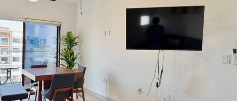 A modern living room with a flat-screen TV mounted on the wall, a dining table with chairs, a ceiling fan, and an air conditioning unit.