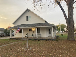 A single-story house with a porch, a flag, and a small tree.