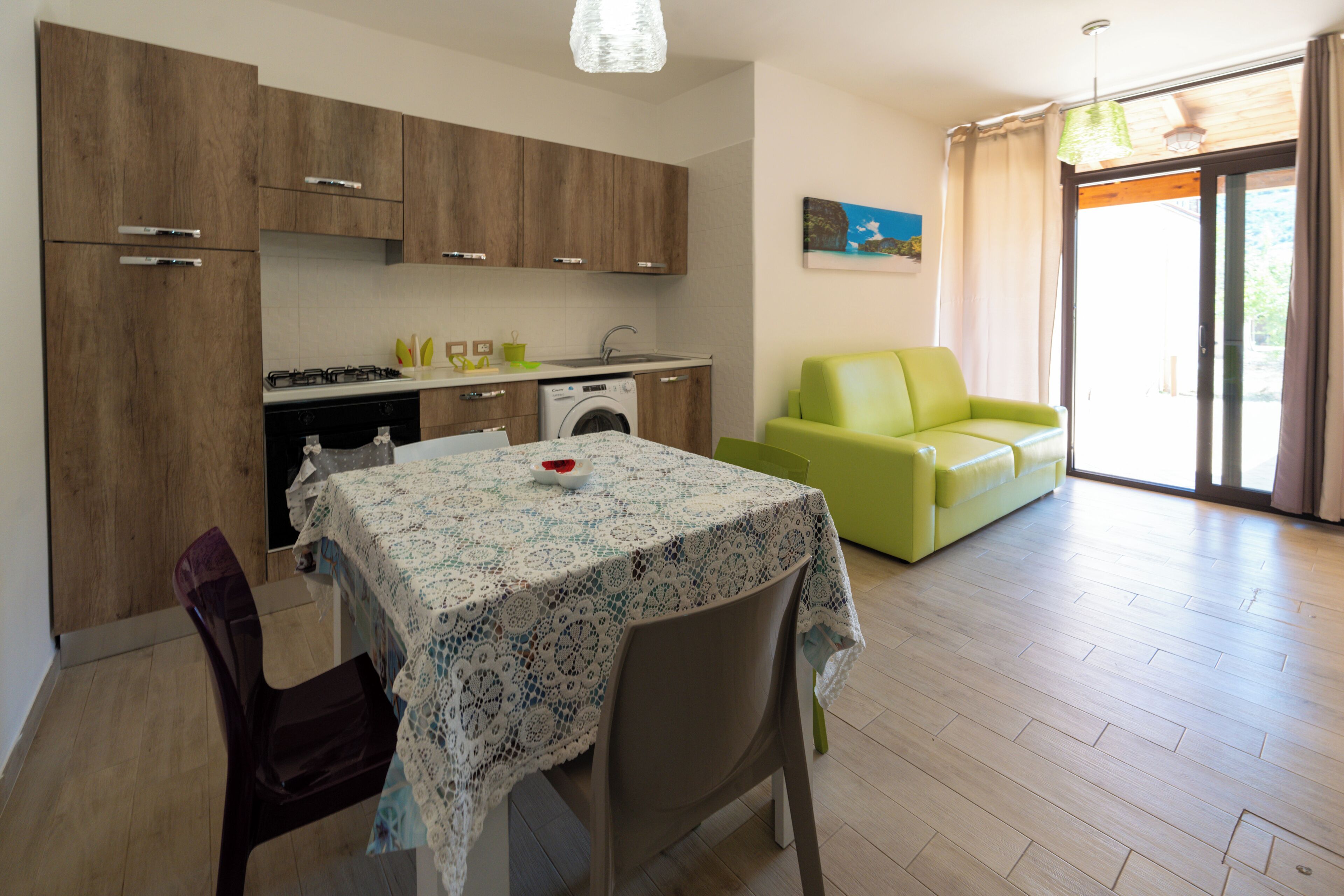 Eine moderne Küche mit Holzausstattung, einem Esstisch mit Spitze und einem grünen Sofa in einem hellen Wohnbereich mit großem Fenster.