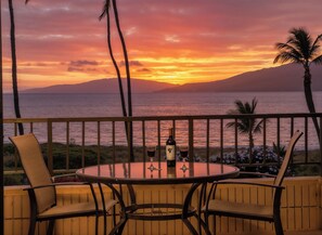 Une table ronde entourée de chaises, placée sur un balcon d’où l’on peut admirer le coucher de soleil sur l’océan. Une bouteille de vin et un verre sont posés sur la table.