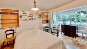 A bedroom with a large bed, wooden furniture, a desk, and a view of greenery through large windows.