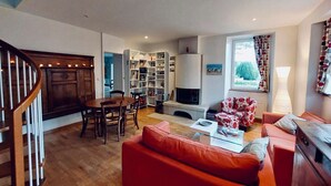 A spacious living area with a wooden staircase, a dining table with chairs, a red sofa, a fireplace, and a bookshelf.