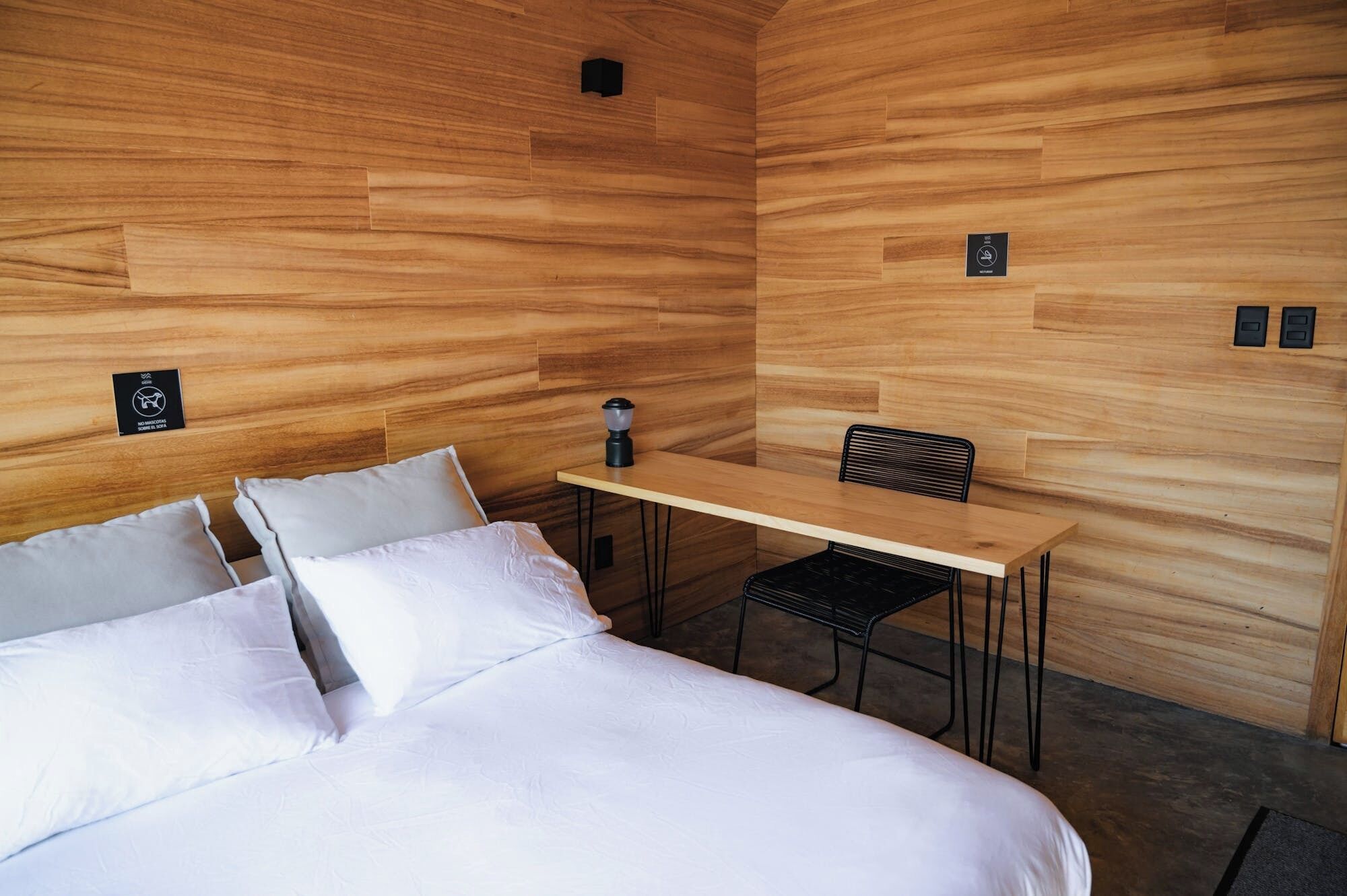 Una habitación moderna con una cama, un escritorio y una silla. Hay carteles en la pared que indican que no se permite fumar ni alimentos del exterior.