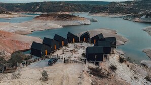 Une série de cabanes aux couleurs sombres, dotées de portes orange, disposées en demi-cercle autour d’un espace central recouvert de gravier, avec un plan d’eau et des collines en arrière-plan.
