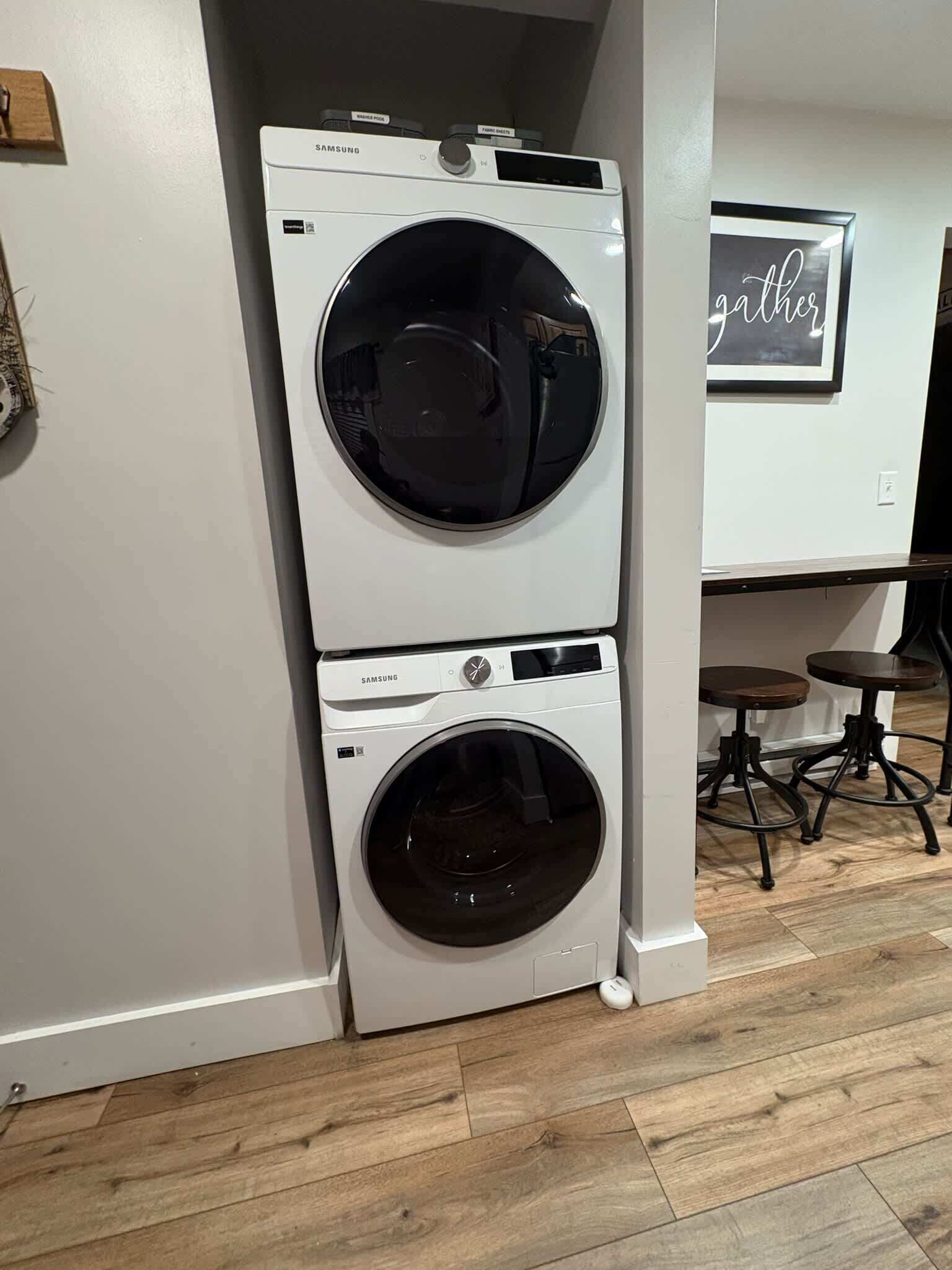 A laundry room with two white Samsung front-loading washers and a wall-mounted sign that says 'gathers.'