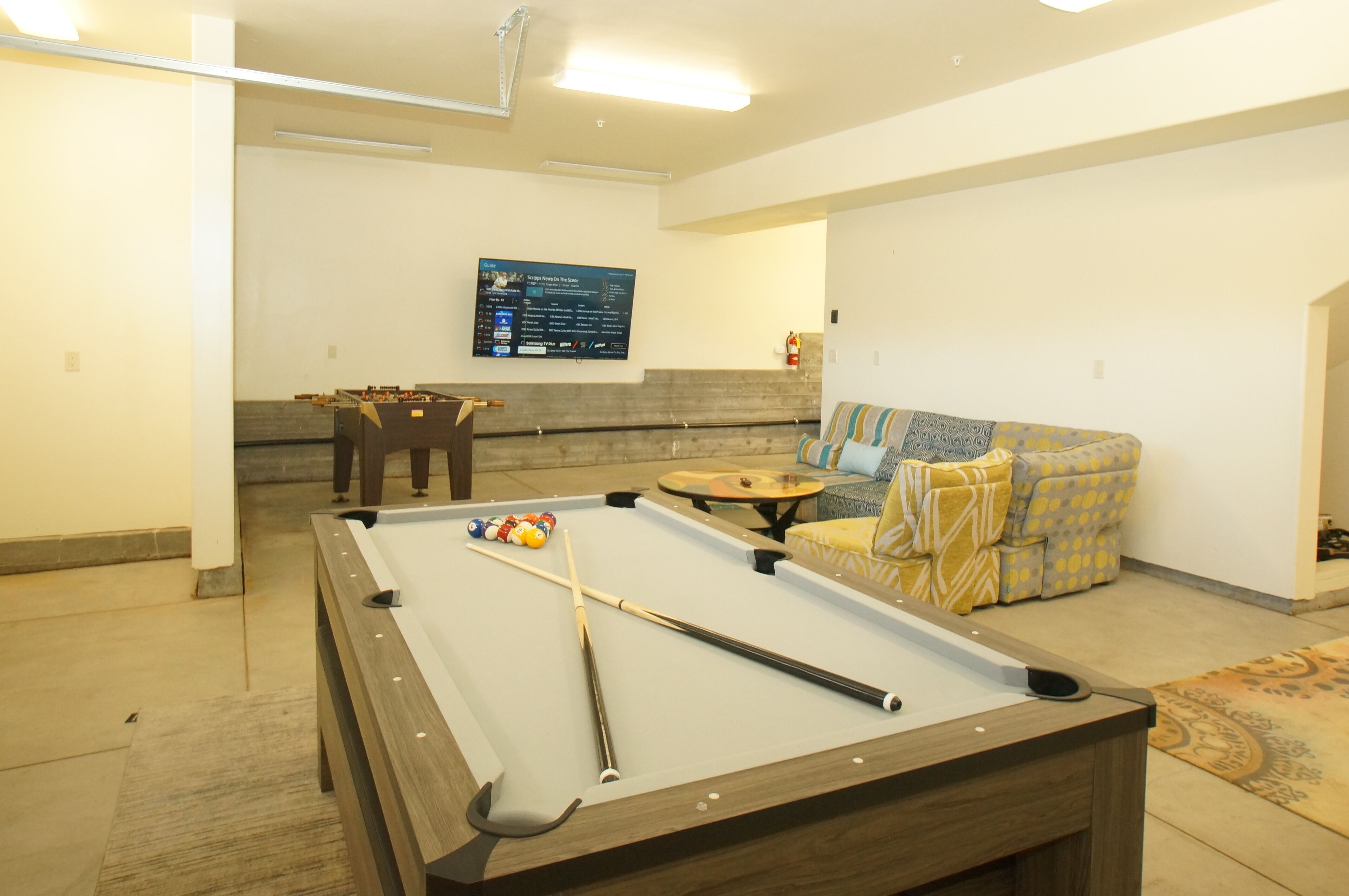 A pool table with balls and cues, a sofa with patterned cushions, and a mounted television in a room with neutral decor.