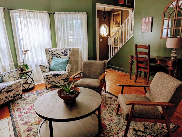 A living room with a round table, two armchairs, a floral armchair, a wooden chair, and a staircase leading to another room.