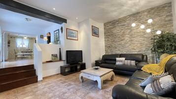 A modern living room with a black leather sofa, a wooden coffee table, and a flat-screen TV. There is a staircase leading to an upper level, and a stone wall with a textured finish.