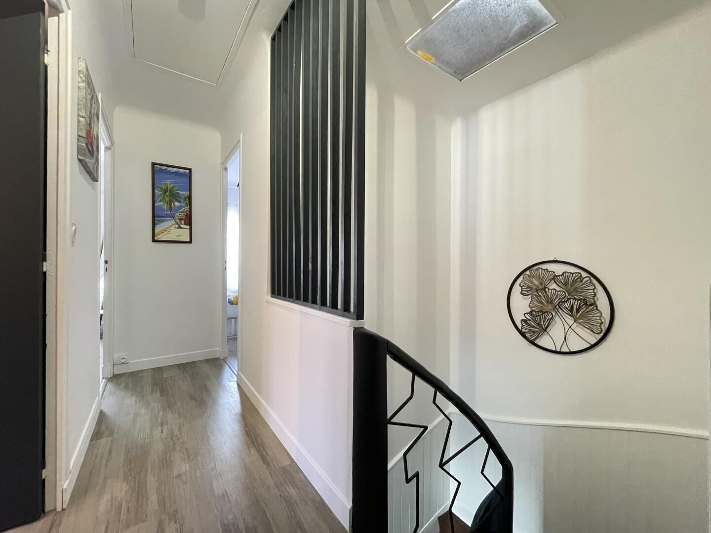 A hallway with a wooden floor, a black railing with a zigzag design, a window with vertical blinds, and a wall-mounted decorative metal piece.
