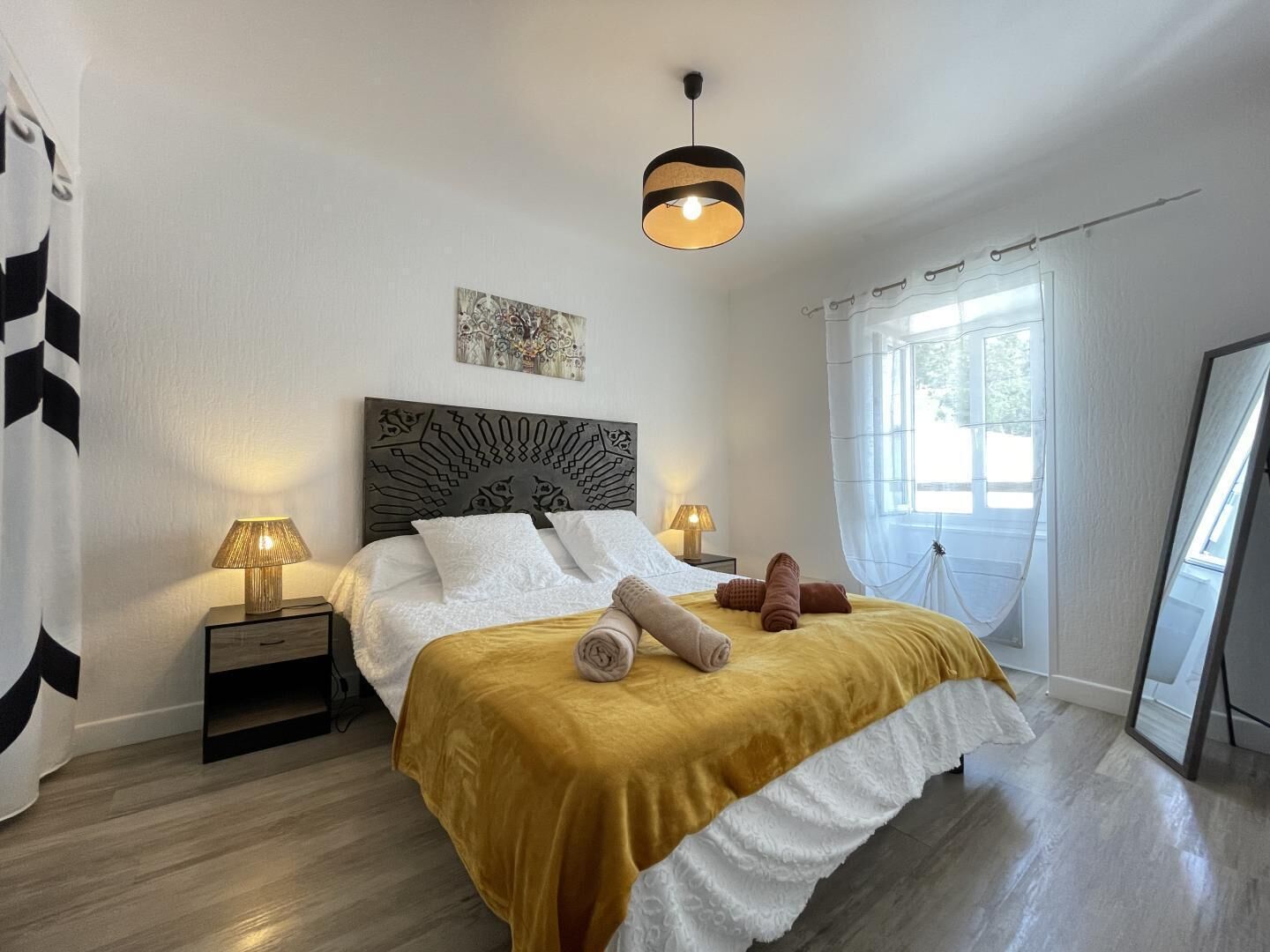 A bedroom with a bed, two bedside tables, a hanging light fixture, a window with curtains, and a wall art piece.