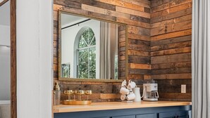 A bathroom with a wooden wall, a large mirror, and a dark blue cabinet. - Downtown Wimberley Treehouse | Hot Tub Under Stars (Wimberley)