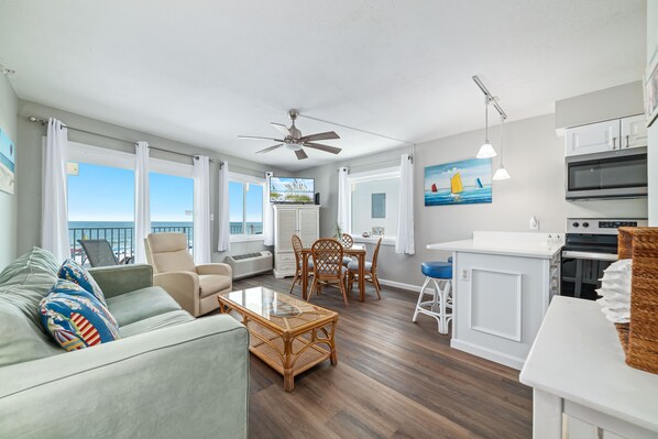 A spacious living room with a sofa, a dining table, and chairs, a kitchen area with an oven and microwave, and large windows with a view of the sea.