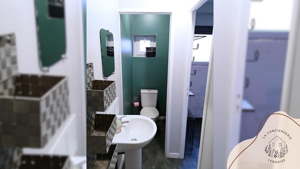 Kylpyhuoneessa on valkoinen pesuallas, wc-istuin ja vihreä seinä. WC-istuimen yläpuolella on ikkuna ja seinällä peili.