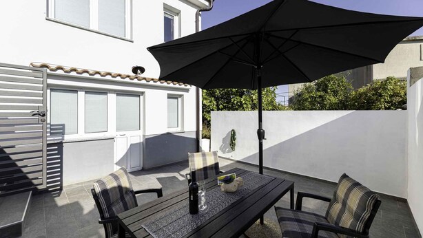 An outdoor patio with a table and chairs, an umbrella, and a white building in the background. - Holiday apartment with air conditioning and terrace (Baška)