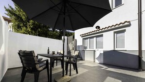 Een terras buiten met een tafel en stoelen, een grote parasol, en een wit gebouw met ramen en een pannendak.