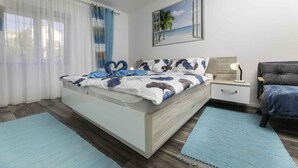 A modern bedroom with a bed, a nightstand, a lamp, and a view of the beach through a window. - Holiday apartment with air conditioning and terrace (Baška)