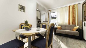 Een knus woongedeelte met een eethoek, bestaande uit een ronde tafel, stoelen en een schaal met fruit. Een bank en een klein tafeltje met een kopje staan bij een raam met gordijnen.
