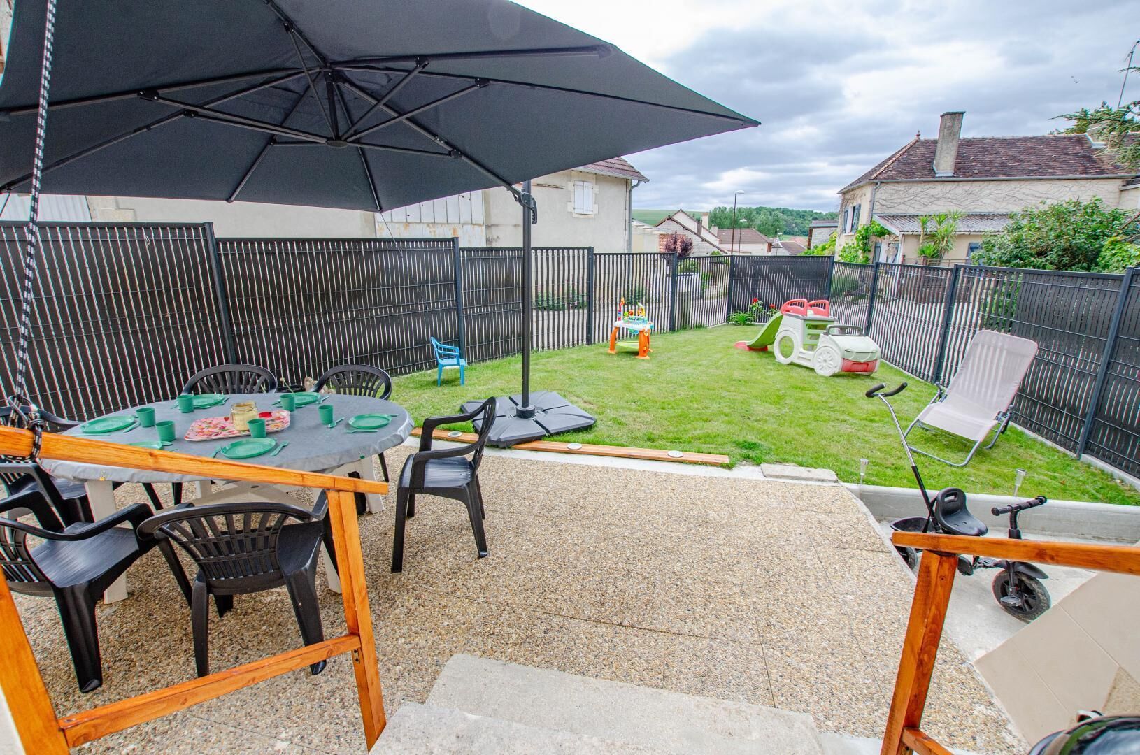 En udendørs terrasse med et bord dækket til fire personer, omgivet af stole, under en stor parasol.