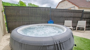 A hot tub in a backyard with a wooden fence, a house in the background, and two white chairs.