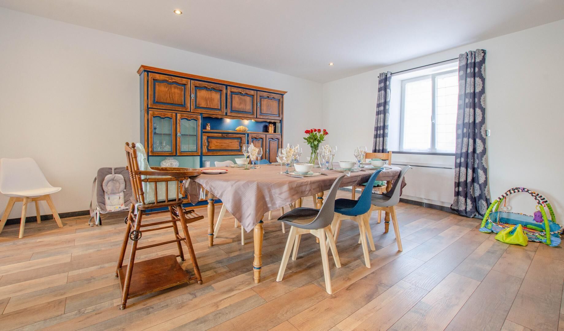 Un comedor con una mesa de madera dispuesta para cuatro personas, rodeada de sillas. Se observa un armario con puertas de cristal y una ventana con cortinas.
