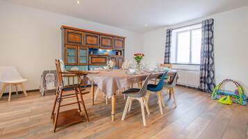 Un comedor con una mesa de madera dispuesta para cuatro personas, rodeada de sillas. Se observa un armario con puertas de cristal y una ventana con cortinas.