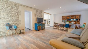 A spacious living area with a stone wall, wooden flooring, a sofa, a TV stand, and a dining area with wooden cabinets and chairs.