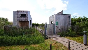 A rustic wooden building with a small window and a door, surrounded by a wooden fence and a pathway leading to it.