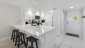 A modern kitchen with white cabinetry, a central island with stools, a microwave, and a sink. - Beachside Ground Floor Condo | Steps to the Beach, Heated Pool & Private Patio 🌴 (Cocoa Beach)