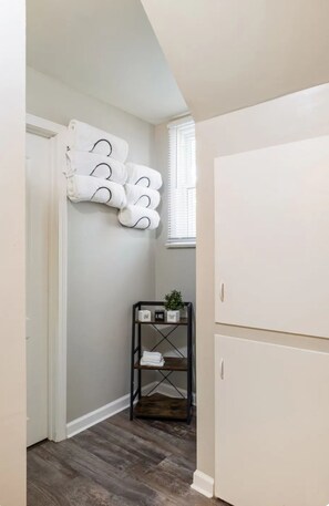 Un pasillo con una puerta blanca, suelo de madera, un armario blanco y una estantería con toallas dobladas y una planta en maceta.