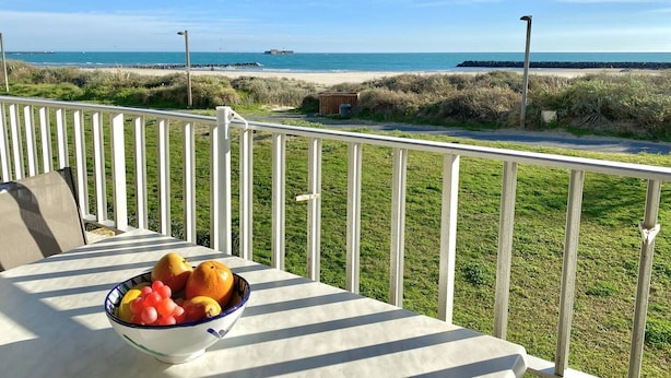 A balcony with a table holding a bowl of fruit, overlooking a beach and dunes.