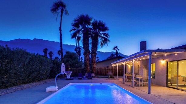 A swimming pool with a basketball hoop, surrounded by palm trees and a house with outdoor seating. - Poolside Living Home (Palm Springs)