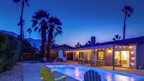 A backyard with a swimming pool, two Adirondack chairs, a basketball hoop, and a patio area with string lights. - Poolside Living Home (Palm Springs)