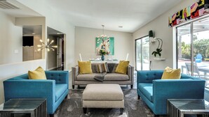 A modern living room with two blue sofas, a grey sofa, and a beige ottoman. There is a chandelier, a painting, and a plant on the wall. - Poolside Living Home (Palm Springs)