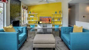 A modern living room with teal sofas, a beige ottoman, and a large flat-screen TV on a white stand. - Poolside Living Home (Palm Springs)