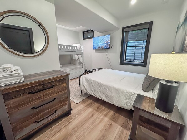 Un dormitorio con literas, un televisor de pantalla plana, una ventana con persianas, un aparador de madera y una lámpara de noche.