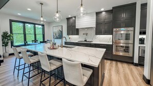 Une cuisine moderne dotée d’un grand îlot central, de meubles de couleur sombre, d’appareils électroménagers en acier inoxydable et de luminaires suspendus.
