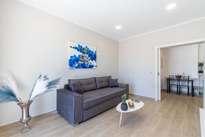 Ein modernes Wohnzimmer mit einem grauen Sofa, einem kleinen runden Tisch mit einer Pflanze, einer Vase mit Federn und einem Buddha-Gemälde an der Wand.