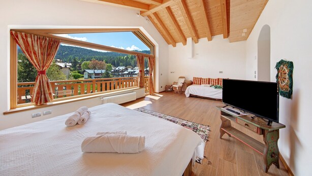 A bedroom with a large window, wooden ceiling beams, a bed with towels, a TV on a wooden stand, and a chair. - Affittacamere Nene – Attic Room with Mountain View and Terrace (Pieve di Cadore)