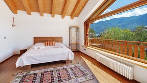 A bedroom with a wooden ceiling, a large window with a view of mountains, a bed with white linens, and a patterned rug. - Affittacamere Nene – Attic Room with Mountain View and Terrace (Pieve di Cadore)